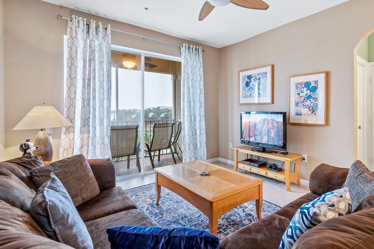 A living room with a sofa set, a coffee table, a TV on a wooden stand, and a balcony with a view.