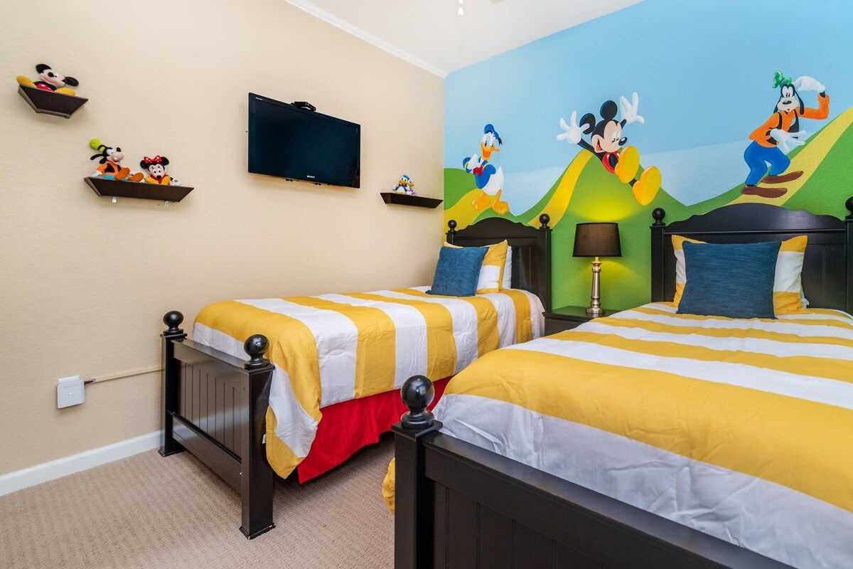 A hotel room with two beds, each with striped bedding, a flat-screen TV on the wall, and a mural of animated characters.