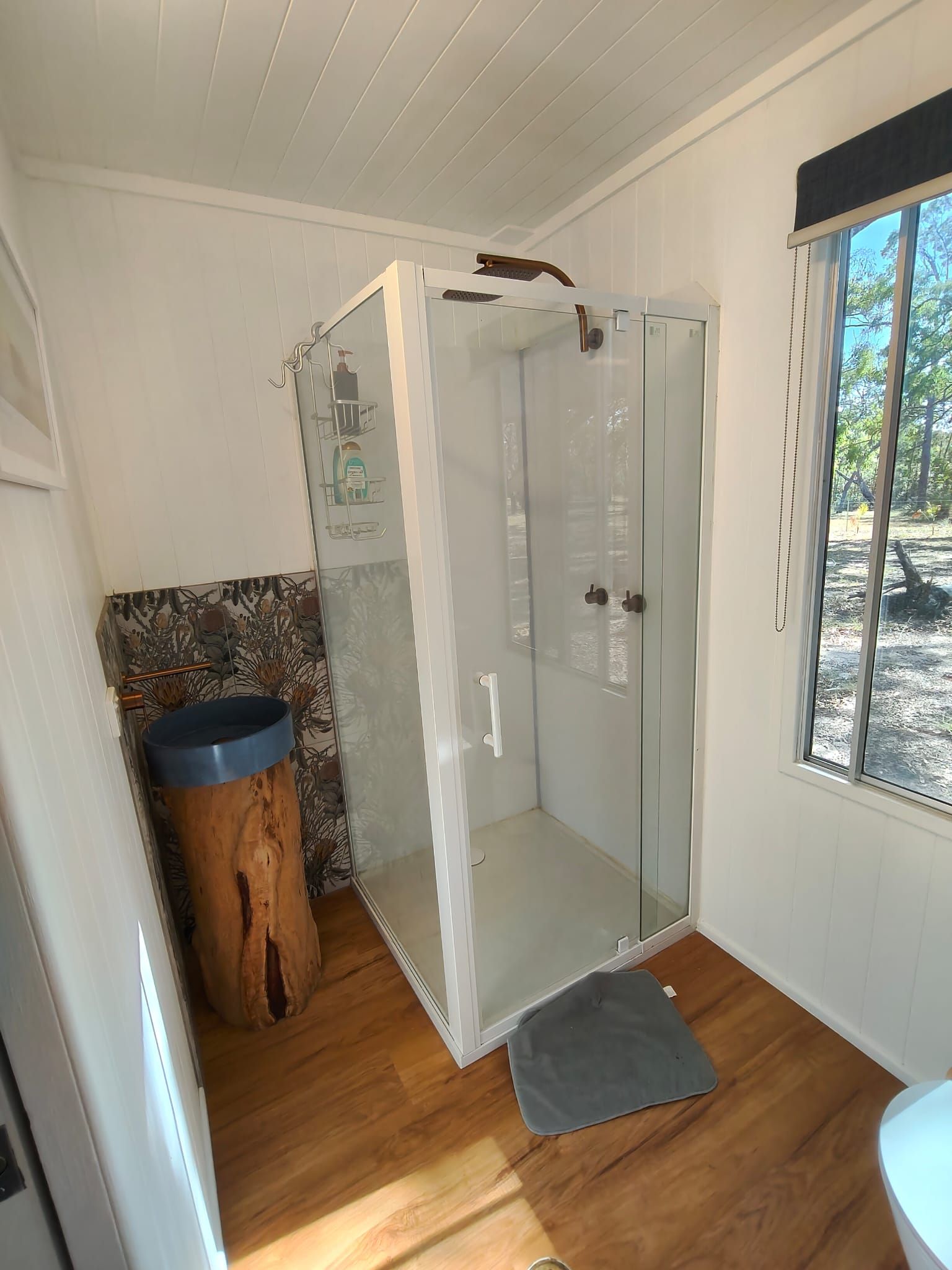 A shower with glass doors, a wooden bench, and a window with a view of trees.