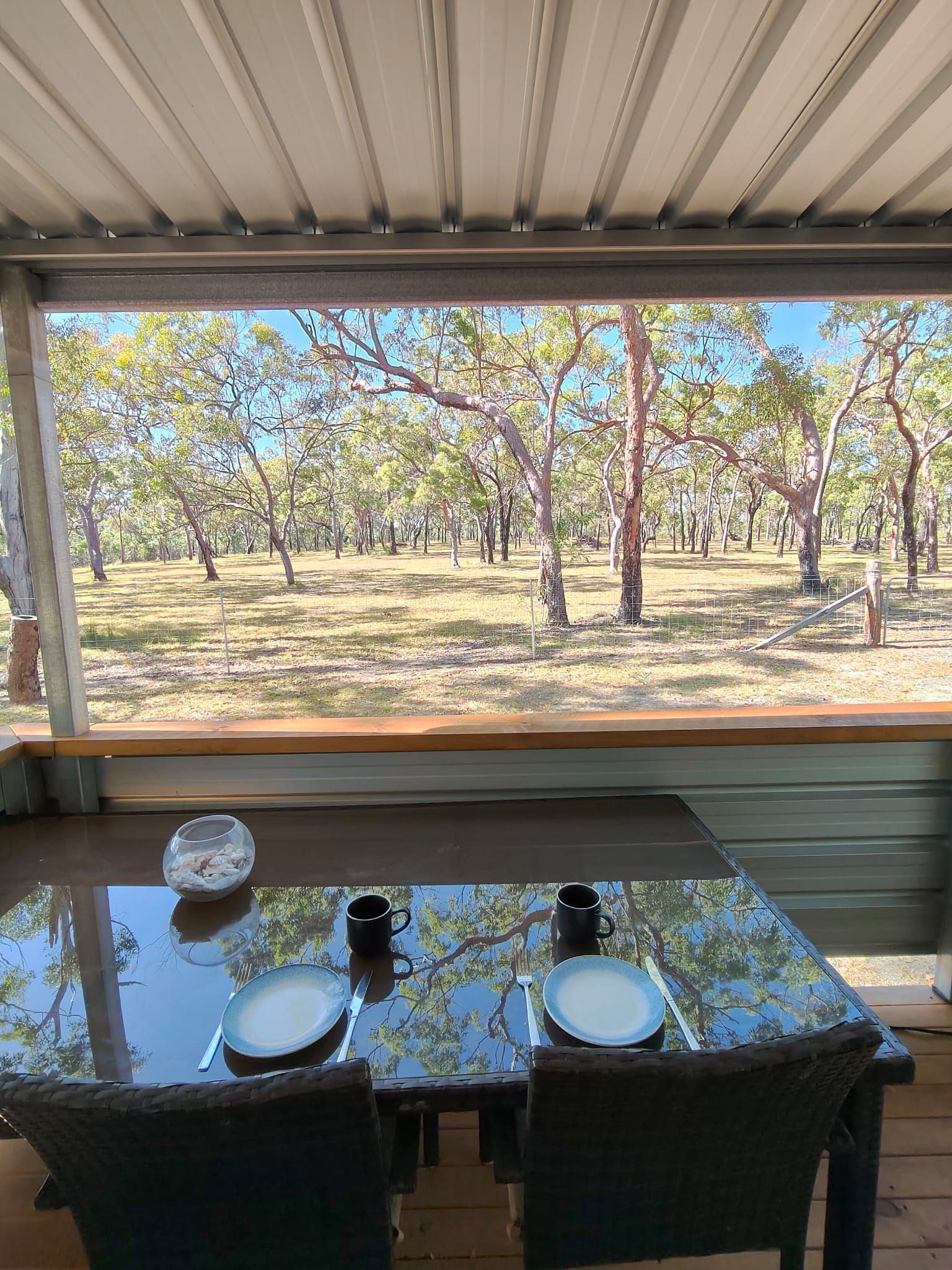 Une table à manger avec deux chaises, un bol et deux tasses, disposée sous un espace abrité, avec vue sur un paysage bordé d’arbres.
