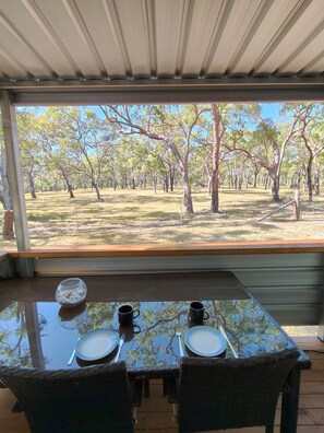 Mesa de comedor con dos sillas, un cuenco y dos tazas, ubicada en un espacio techado con vistas a un paisaje arbolado.