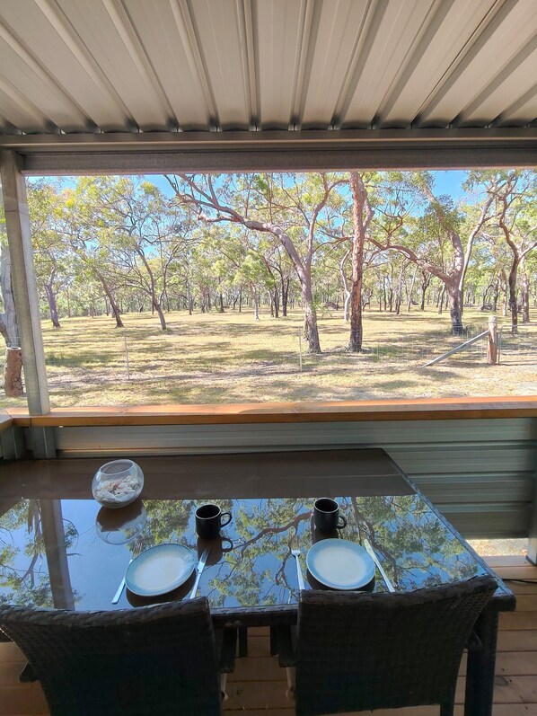 A dining table with two chairs, a bowl, and two cups, set under a covered area with a view of a tree-lined landscape.