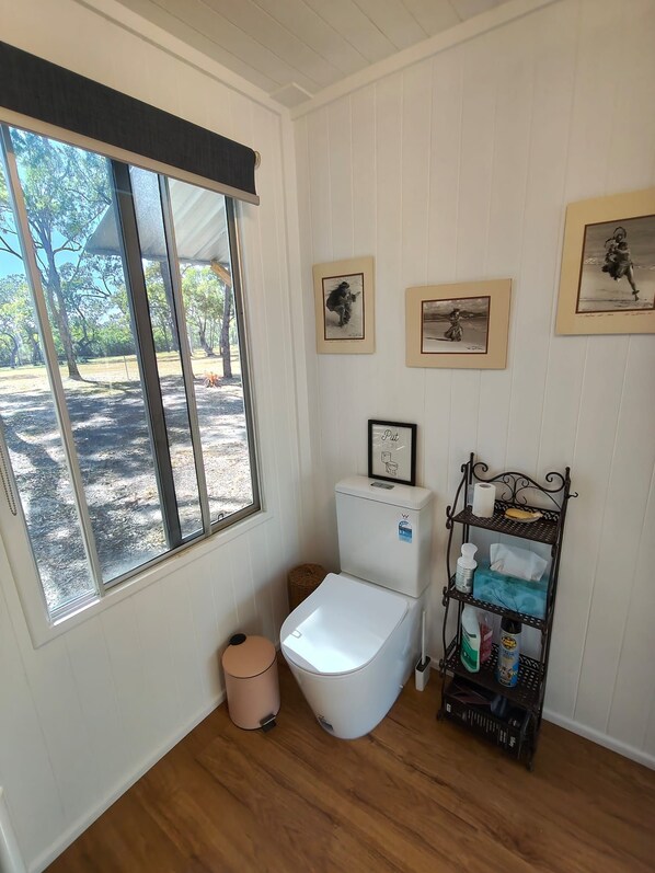 A bathroom with a toilet, a waste bin, a toilet paper holder, and a shelf with toiletries.
