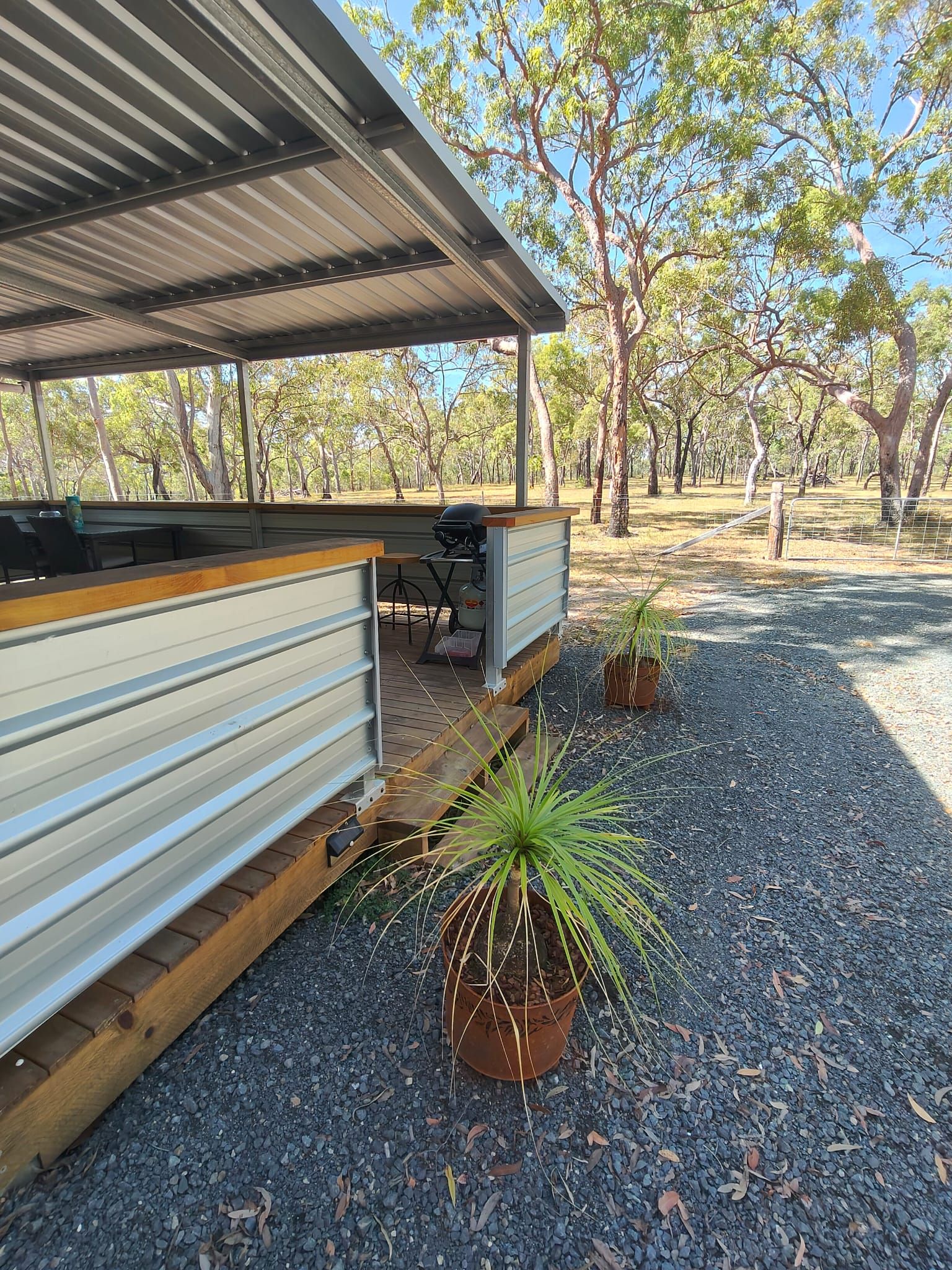 A covered outdoor seating area with a grill, surrounded by trees and gravel.