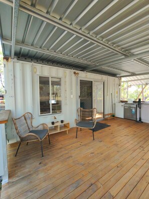 A covered outdoor area with a wooden deck, two wicker chairs, a small table, and a refrigerator.