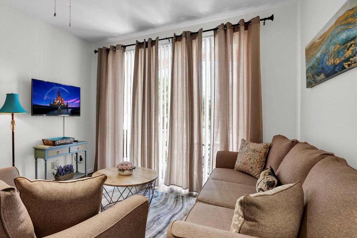 Ein Wohnzimmer mit einer Sofagarnitur, einem Couchtisch, einem an der Wand befestigten Fernseher und einem Fenster mit Vorhängen.