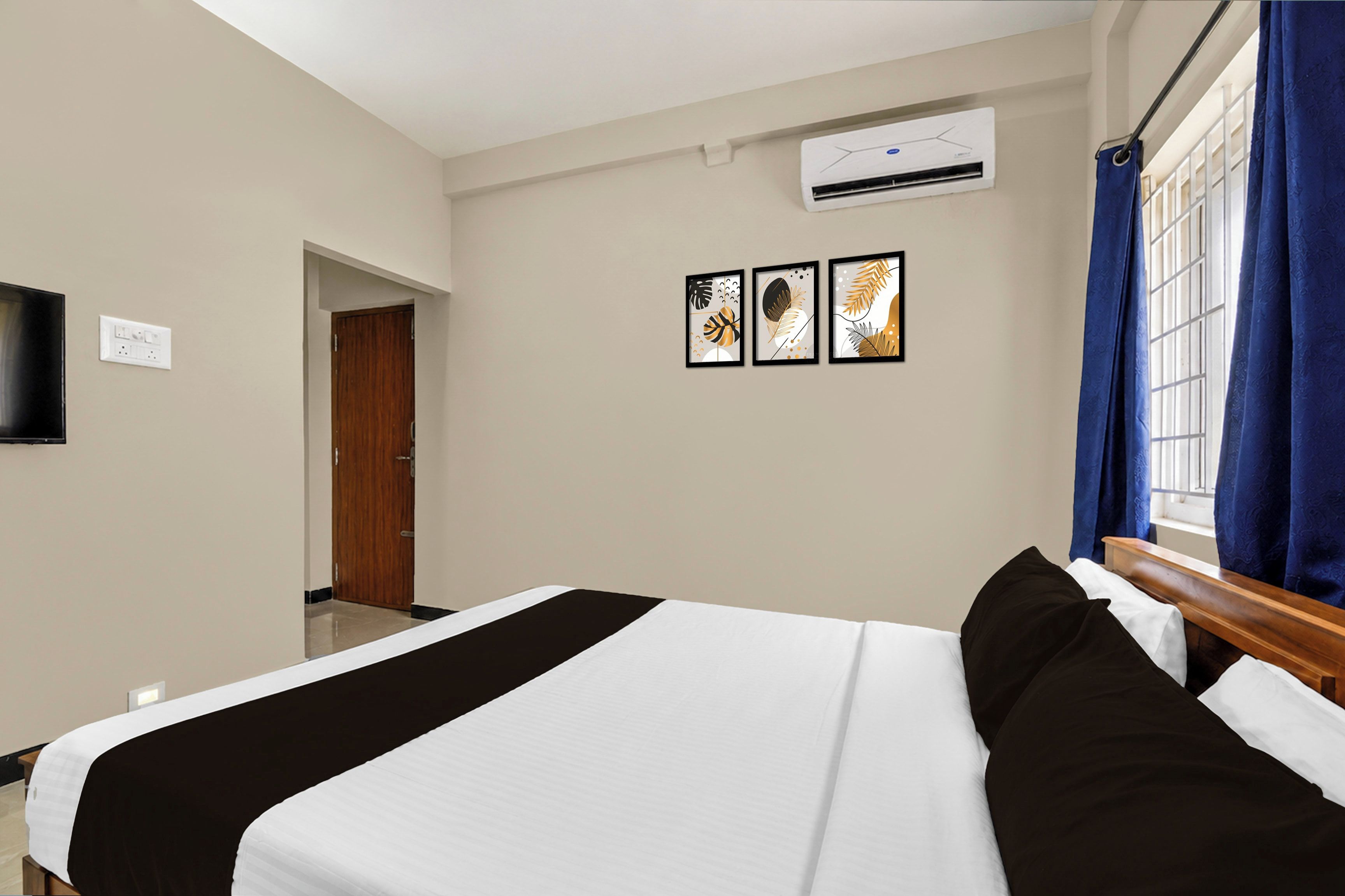 A hotel room with a bed, a flat-screen TV mounted on the wall, an air conditioning unit, and a window with blue curtains.