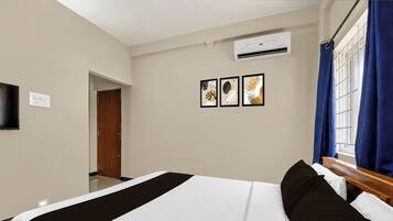A hotel room with a bed, a flat-screen TV mounted on the wall, an air conditioning unit, and a window with blue curtains.