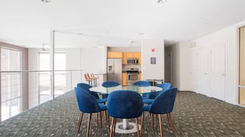 A conference room with a glass table and blue chairs, a kitchenette in the background, and a view of the outside through large windows.