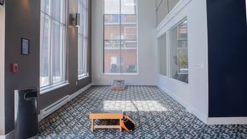 A room with large windows, a fire alarm, a trash bin, and a small wooden table.