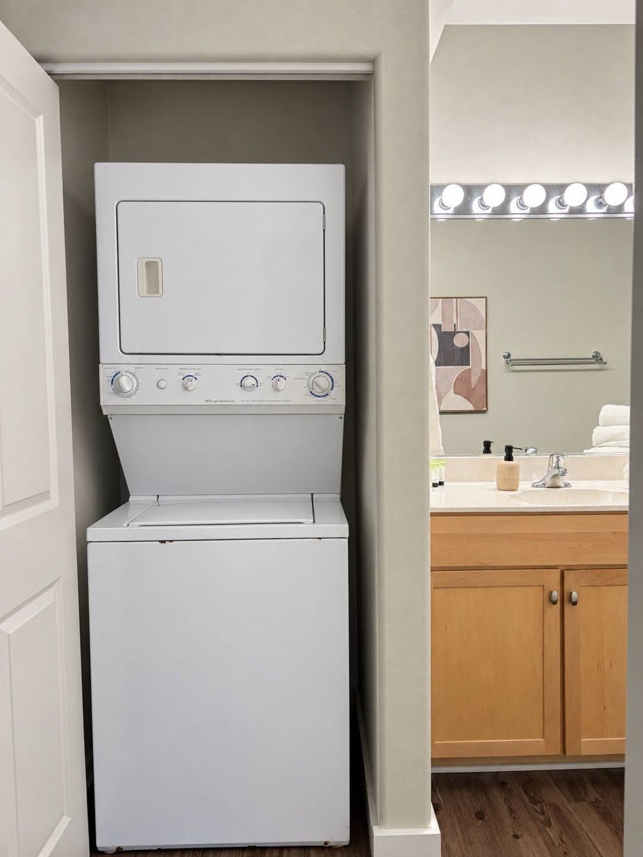 A compact laundry room with a stacked washer and dryer, a wooden cabinet, and a mirror with lights above the sink.