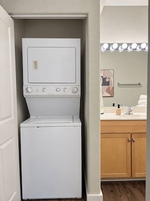 A compact laundry room with a stacked washer and dryer, a wooden cabinet, and a mirror with lights above the sink. - Third Ward 2BR + Den 2-Story Penthouse | Sleeps 10 (Milwaukee)