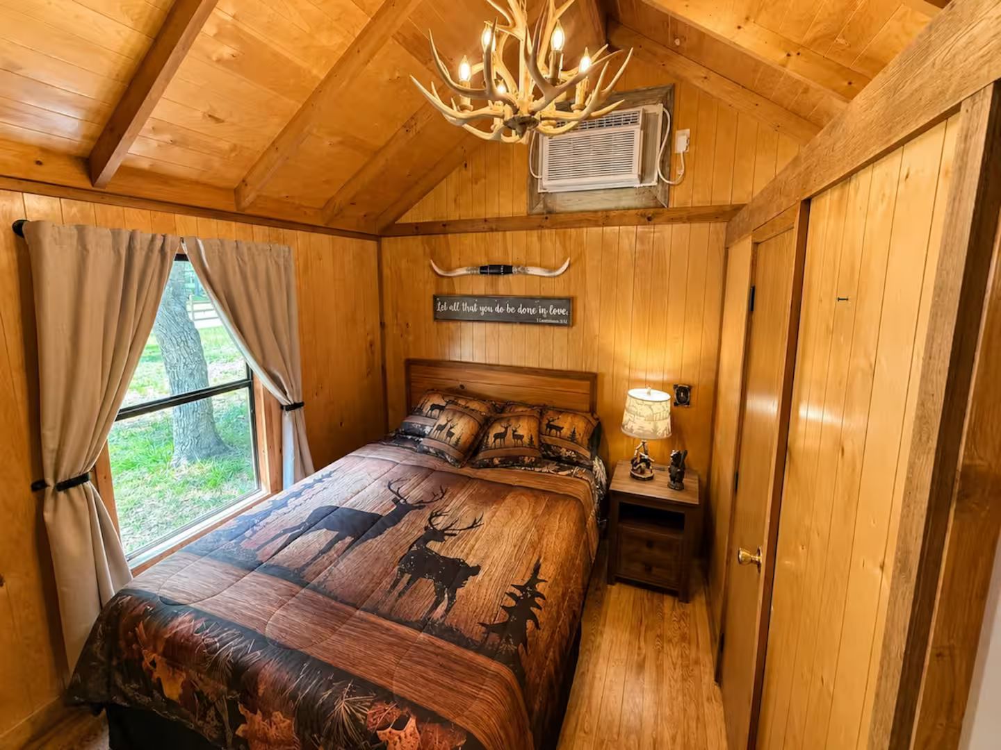 Een slaapkamer in een houten chalet met een bed met patronen van herten en gewei, een kroonluchter en een wandairconditioning.
