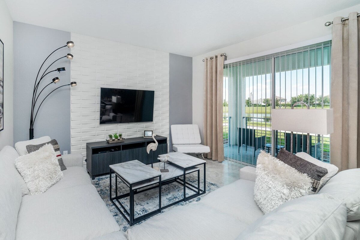 A modern living room with a white sofa, a flat-screen TV, a coffee table, and a balcony with curtains.