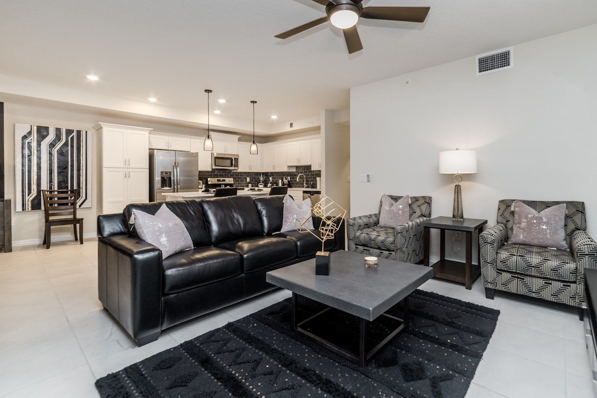 A modern living room with a black leather sofa, patterned armchairs, a coffee table, and a ceiling fan.