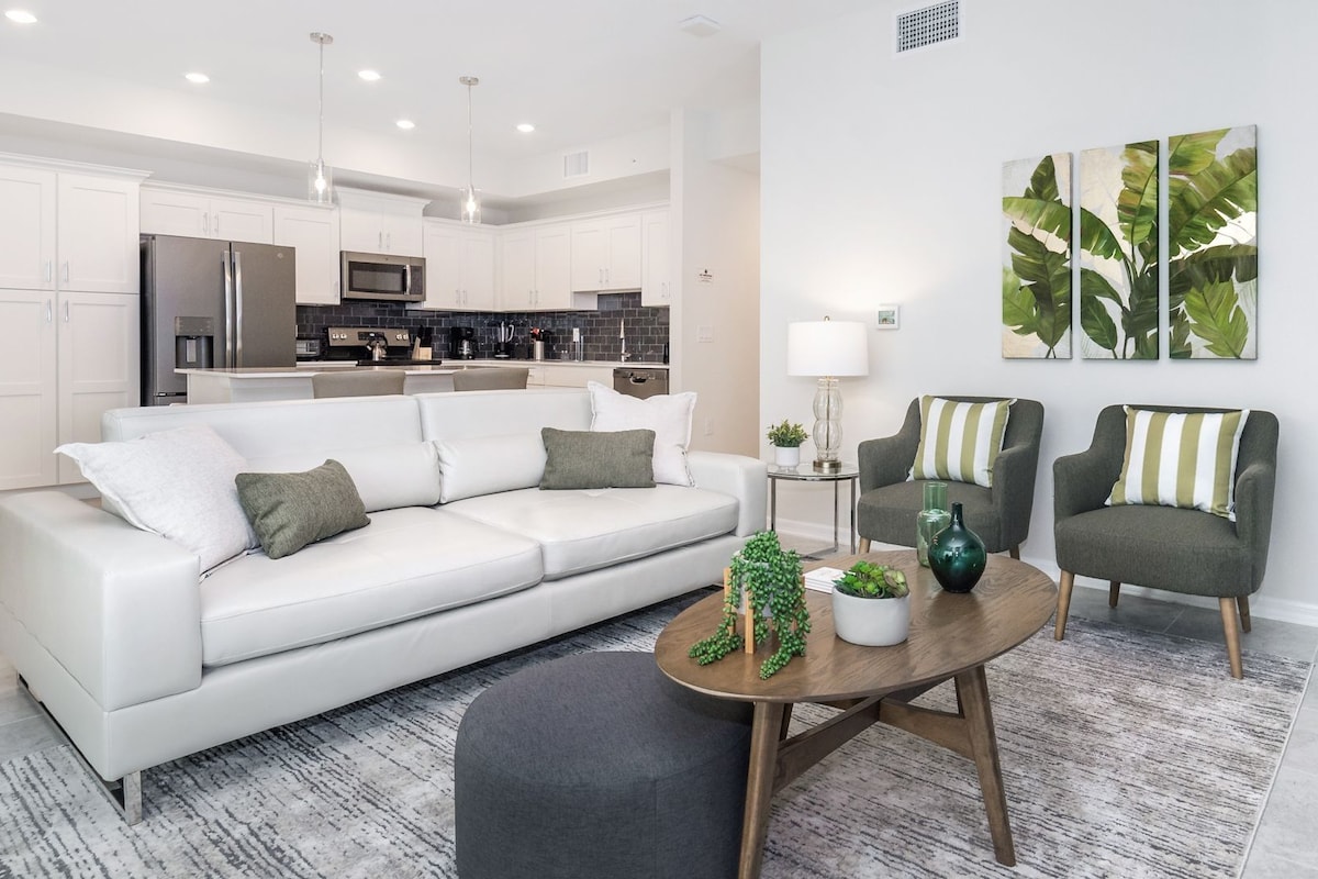 A modern living room with a white sofa, a round coffee table, and two armchairs. There is a kitchen in the background with white cabinets and stainless steel appliances.