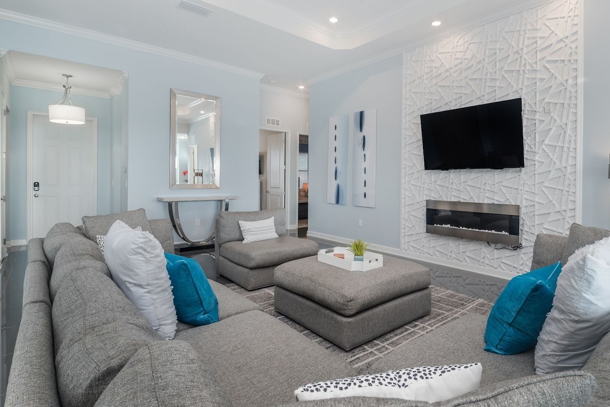 A modern living room with a grey sofa set, a coffee table, a flat-screen TV mounted on a textured wall, and a fireplace.