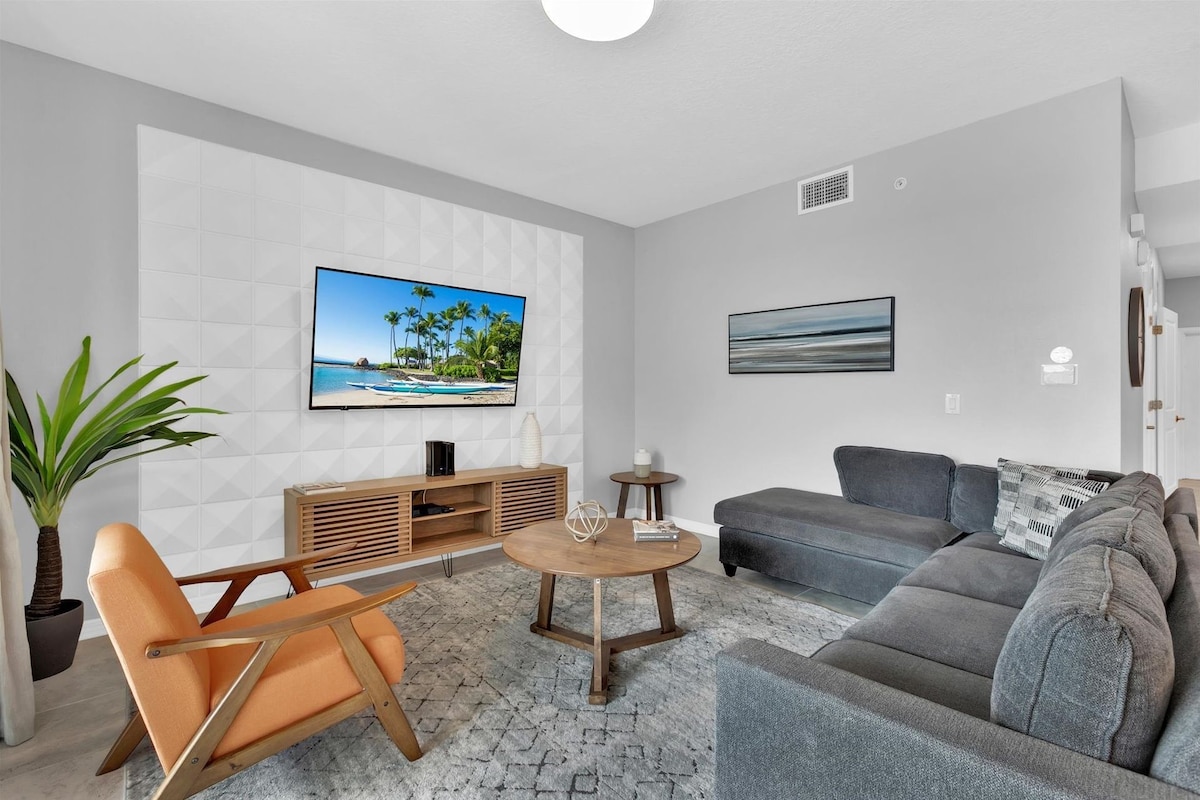 A modern living room with a flat-screen TV, a grey sofa, a wooden coffee table, and a potted plant.