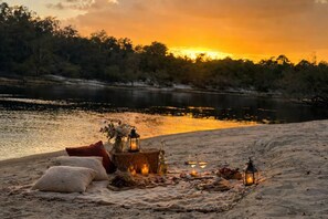 A beach setting with a picnic setup including a basket of fruits, a lantern, and a blanket. - Owl Hollow Cottage - Riverside Escape (Branford)