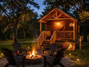 A wooden cabin with a porch, a fire pit, and two Adirondack chairs.