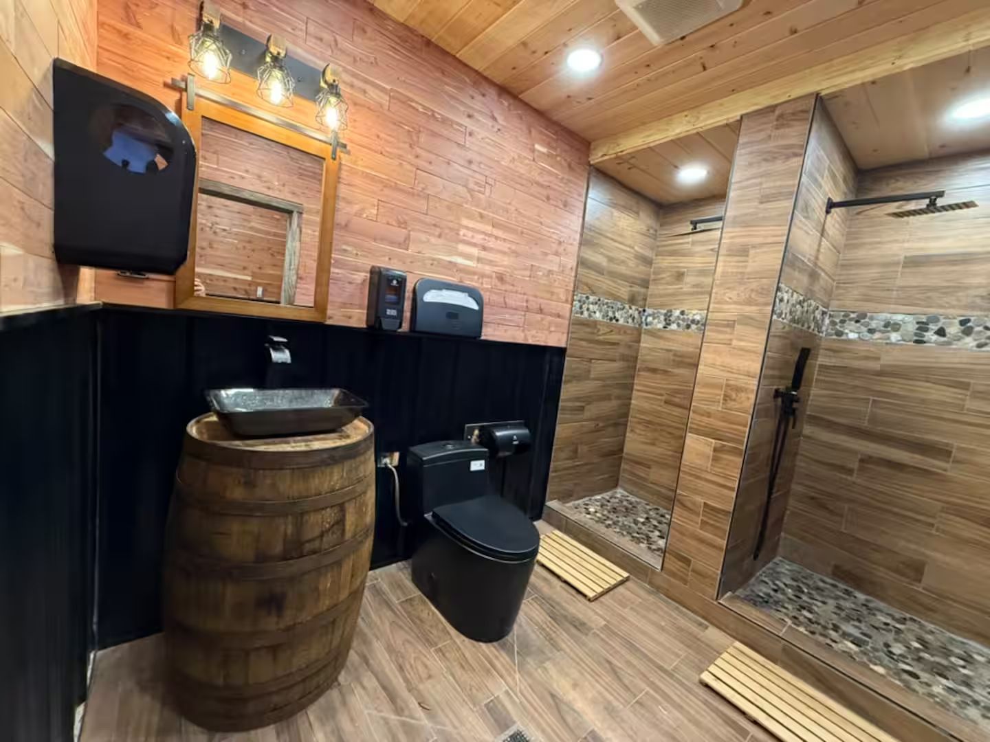 Een badkamer met een wastafel in de vorm van een houten vat, een zwart toilet en een douchegedeelte met houten wanden en een stenen accent.