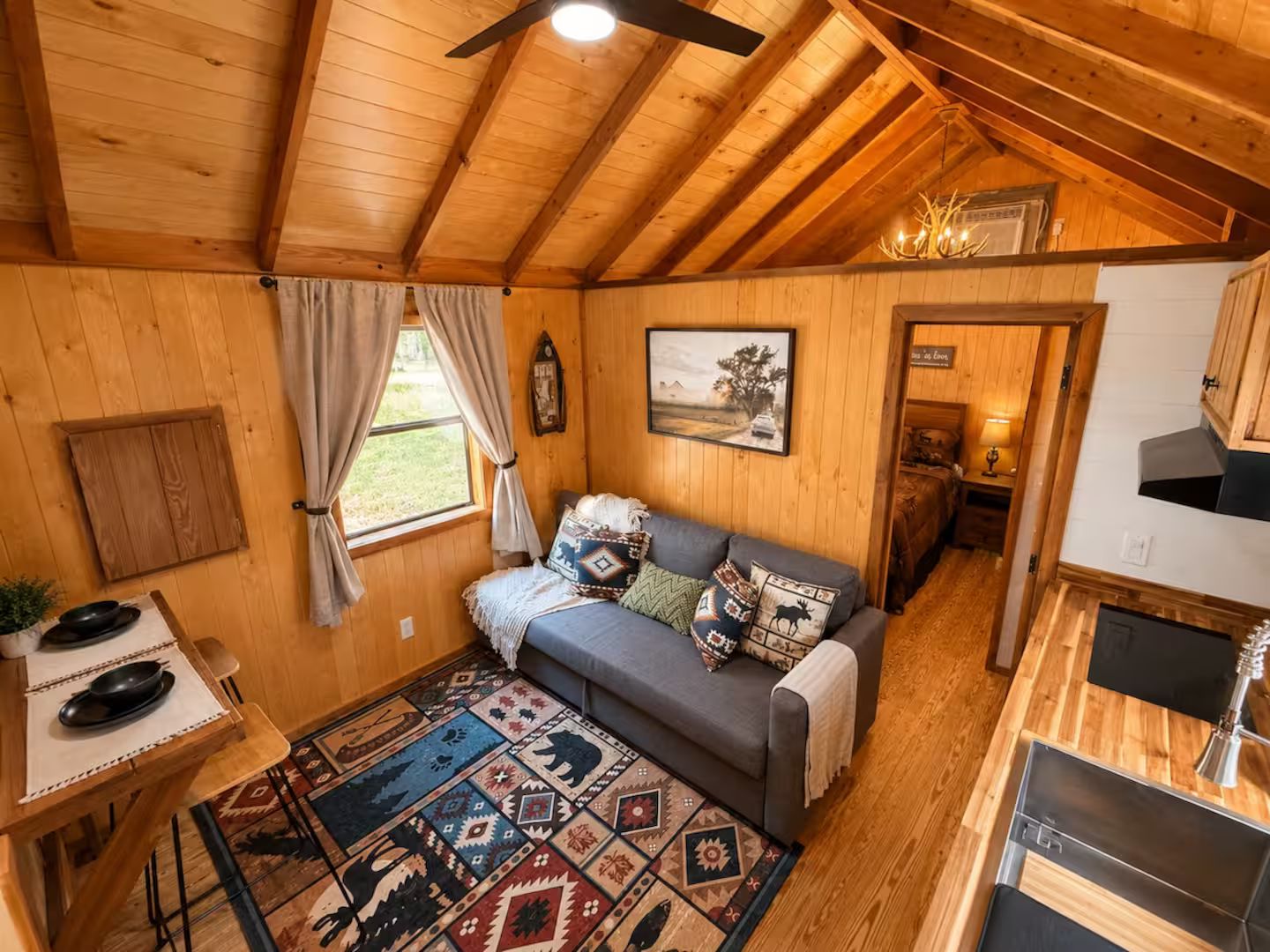 Interior acogedor de una cabaña con techo de madera, un sofá con cojines estampados, una mesa de comedor con un posavasos y una alfombra con diseños geométricos.