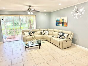 Un salon avec un canapé d’angle beige, une table basse en verre, un ventilateur de plafond et un tableau représentant un paysage urbain accroché au mur.