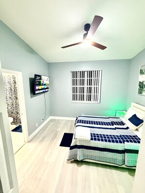 Une chambre à coucher avec un lit, un ventilateur de plafond, un téléviseur fixé au mur et une fenêtre avec des barreaux.