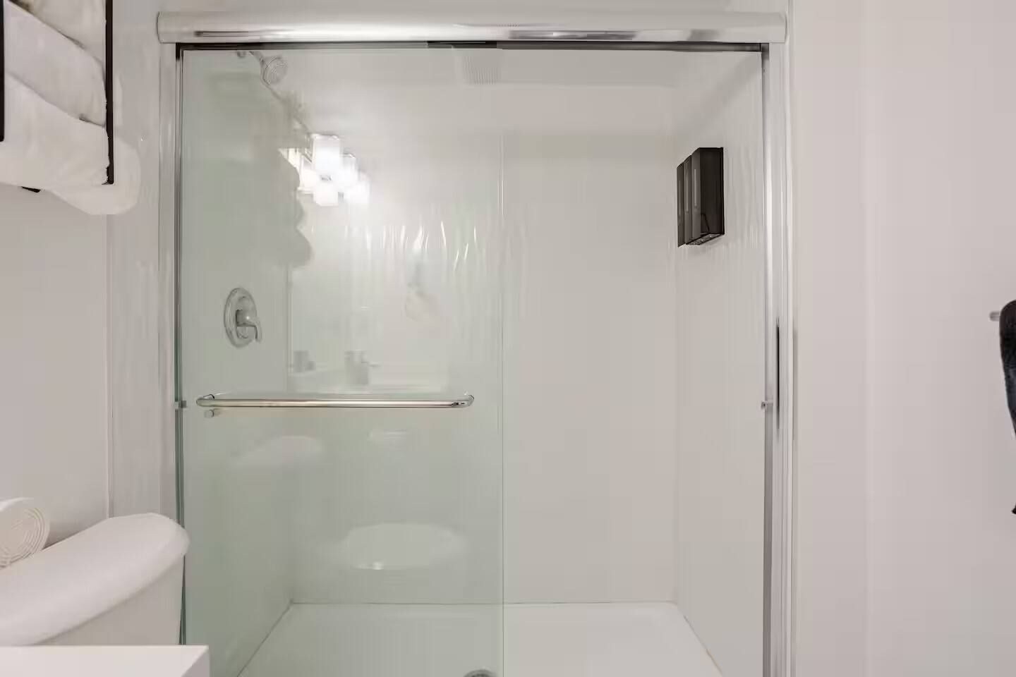 A glass-enclosed shower stall with a handrail, a white sink, and a small black object on the wall.