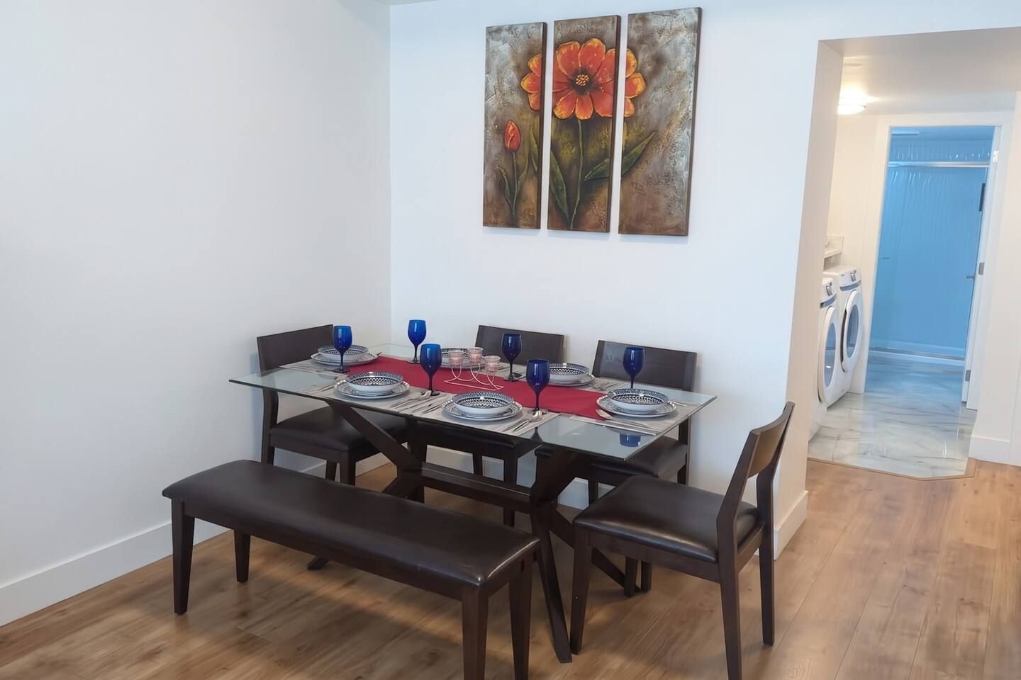 A dining area with a glass table set for four, dark chairs, and a bench. A painting of flowers hangs above the table.