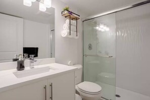 A modern bathroom with a glass shower enclosure, a white sink, and a mirror with lights above.