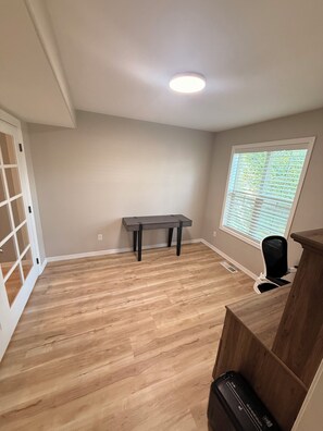 Una habitación con piso de madera, una mesa, un ventanal con persianas y una lámpara en el techo.