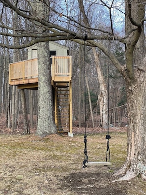 Une cabane perchée dans un arbre, dotée d’une terrasse en bois et d’une échelle, entourée d’arbres dans une zone boisée.