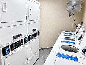 Un área de lavandería con varias lavadoras y secadoras, un ventilador de pared y un panel de control con pantallas digitales.