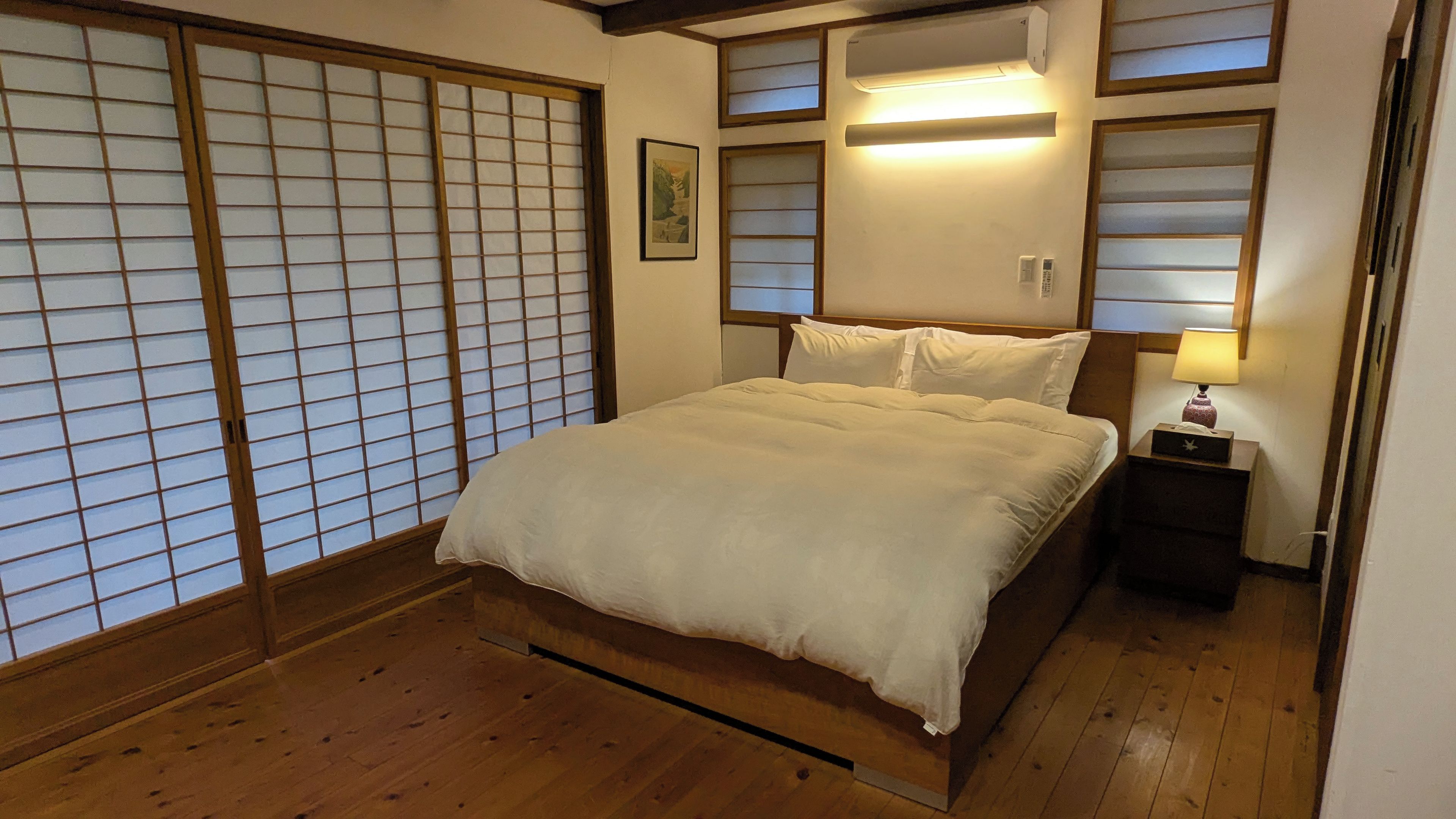 A traditional Japanese-style bedroom with a wooden floor, a bed with white bedding, sliding shoji doors, and a bedside table with a lamp.