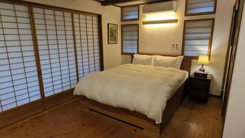 A traditional Japanese-style bedroom with a wooden floor, a bed with white bedding, sliding shoji doors, and a bedside table with a lamp.