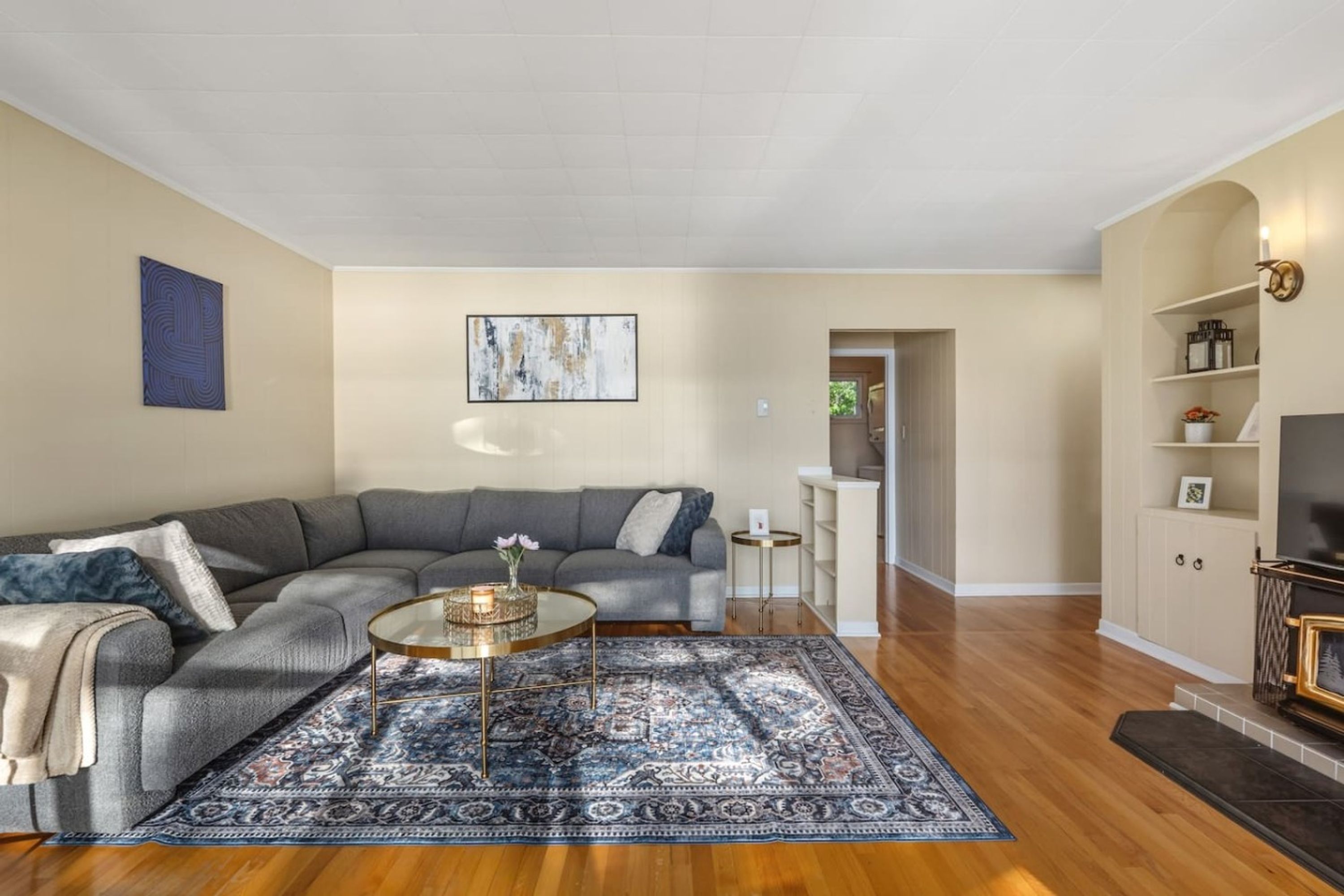 A living room with a grey sectional sofa, a round coffee table, a patterned rug, wooden flooring, a flat-screen TV, and a bookshelf.