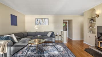 A living room with a grey sectional sofa, a round coffee table, a patterned rug, wooden flooring, a flat-screen TV, and a bookshelf.