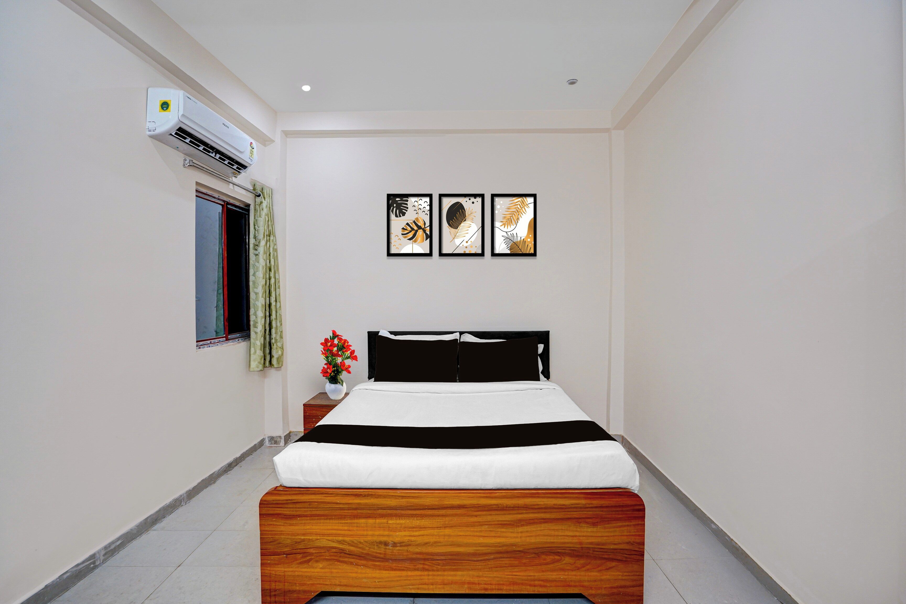 Ein Hotelzimmer mit einem Bett, einem Nachttisch, einem Fenster mit Vorhängen, einer Klimaanlage und drei an der Wand befestigten Kunstwerken.