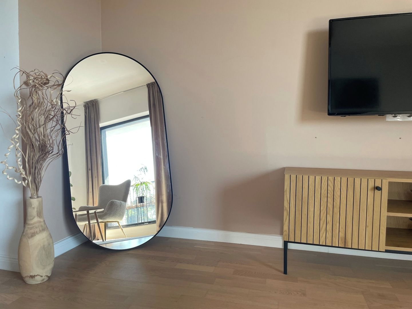 A room with a large oval mirror, a wooden cabinet, and a flat-screen TV mounted on the wall.