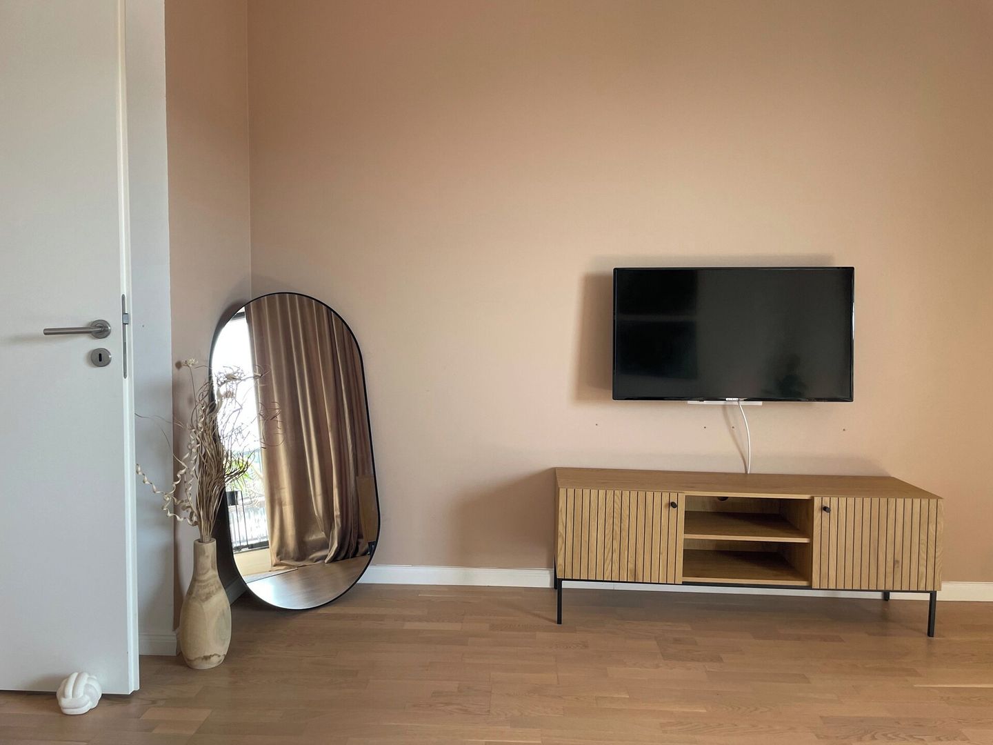 Una habitación con un armario de madera, un televisor de pantalla plana montado en la pared, un espejo redondo apoyado contra la pared y una puerta con cerradura.