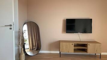 Una habitación con un armario de madera, un televisor de pantalla plana montado en la pared, un espejo redondo apoyado contra la pared y una puerta con cerradura.