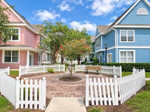 A residential neighborhood with a central circular brick pathway, surrounded by white picket fences, and houses with pastel-colored exteriors. - Storybook House | 4BR Disney Area Retreat (Kissimmee)