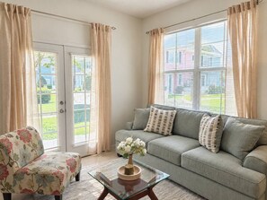 Uma sala de estar com sofá, poltrona floral, mesa de centro de vidro com um vaso e porta de vidro deslizante que dá para um jardim.