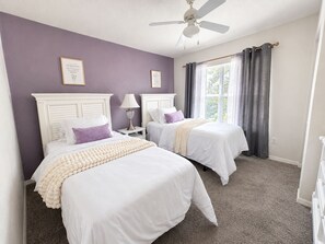 Quarto com duas camas, ventilador de teto e janela com cortinas.
