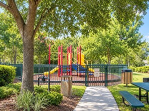 A playground with a slide and climbing structures, surrounded by a green fence and a tree. - Storybook House | 4BR Disney Area Retreat (Kissimmee)