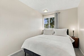 Een slaapkamer met een bed, een nachtkastje met lamp, een raam met gordijnen en een plafondlamp.