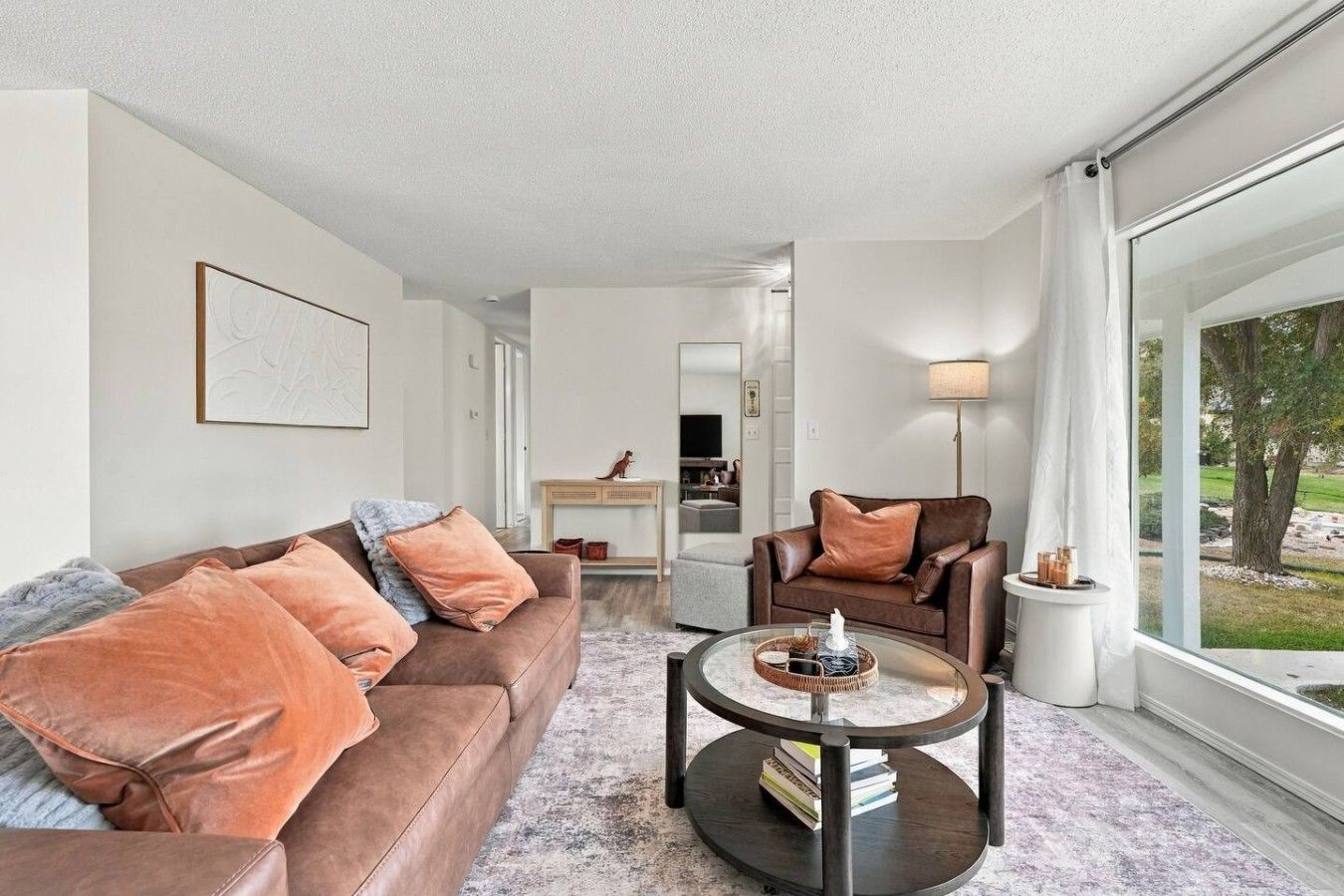 A modern living room with a brown leather sofa, a round coffee table, and a view of a garden through large windows.