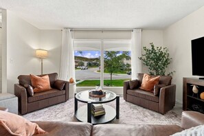 A living room with two sofas, a coffee table, a lamp, a TV stand, and a view of a street and houses.