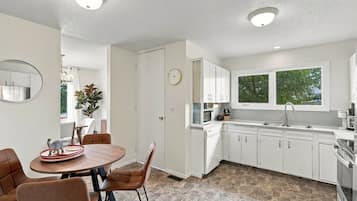 A modern kitchen with white cabinets, a dining area with a round table and chairs, and a window providing a view of greenery outside.